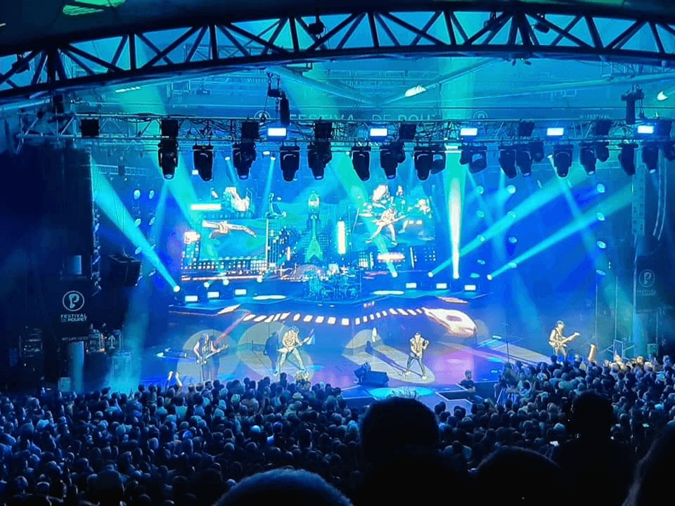 Crowd facing a covered concert stage with blue lighting and a large screen backdrop.