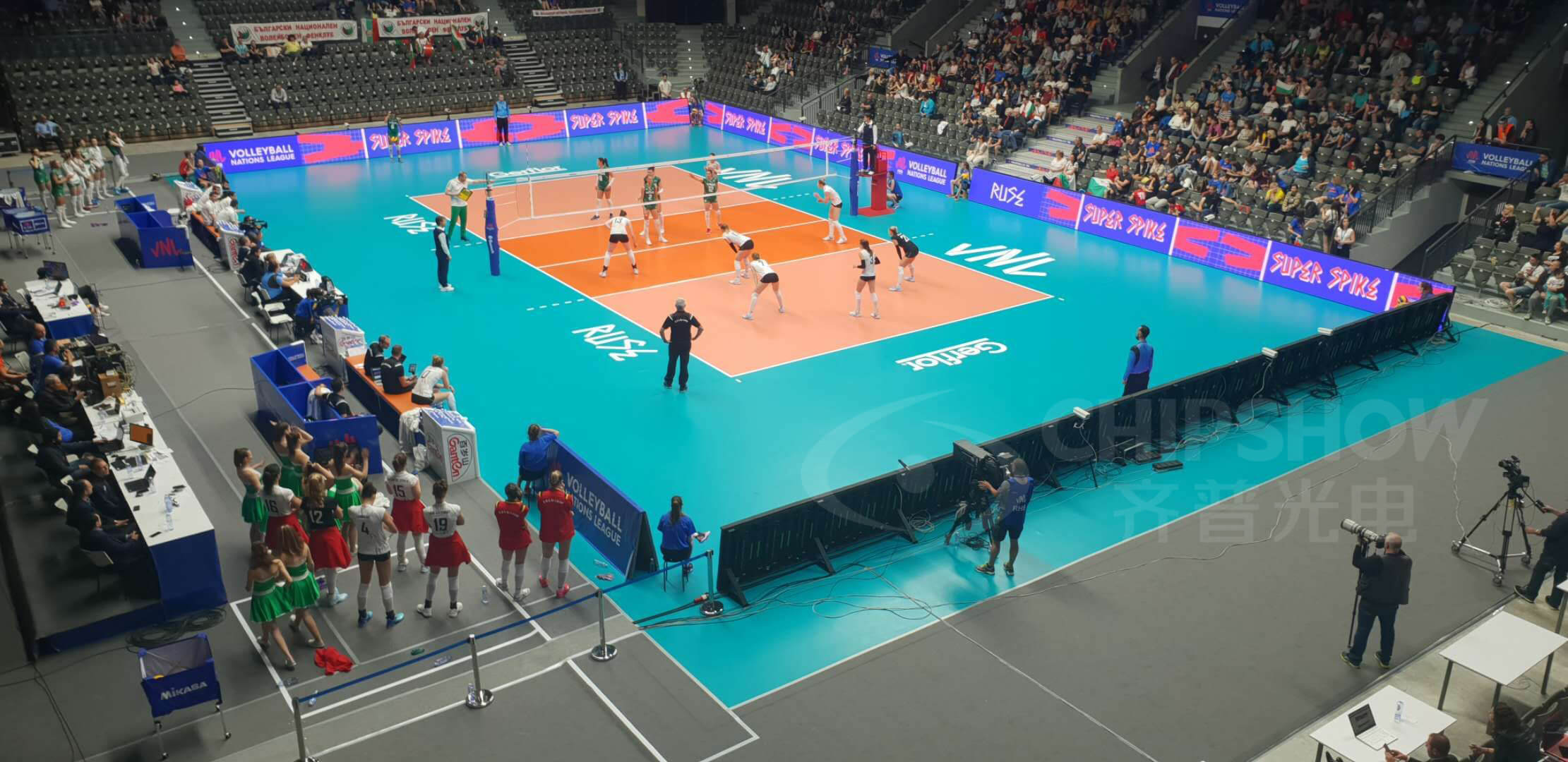 Indoor volleyball match with illuminated advertising panels along the court edges.