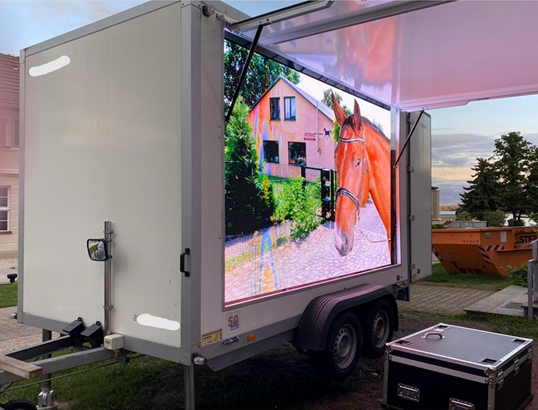 Screen showing a horse beside a house, mounted inside a trailer under an open side canopy.