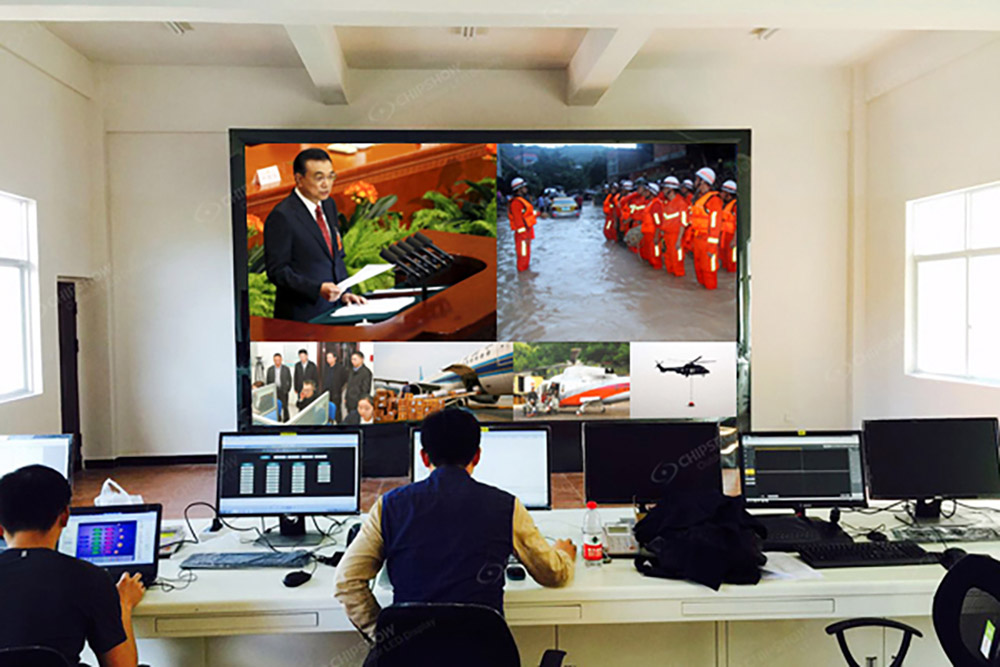 People at computer desks facing a wall screen divided into several video windows.