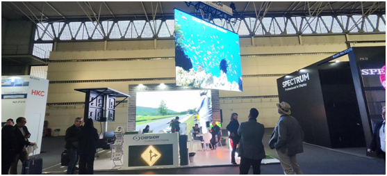 Exhibition stand with a suspended screen, lower displays and visitors in the aisle.
