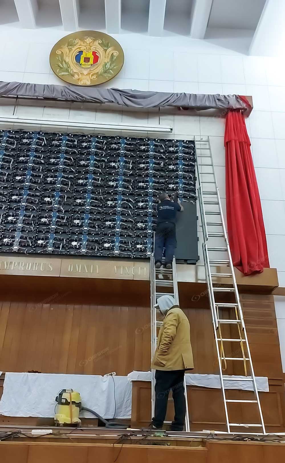 Worker beside a ladder below an exposed display-panel grid above a wooden stage.