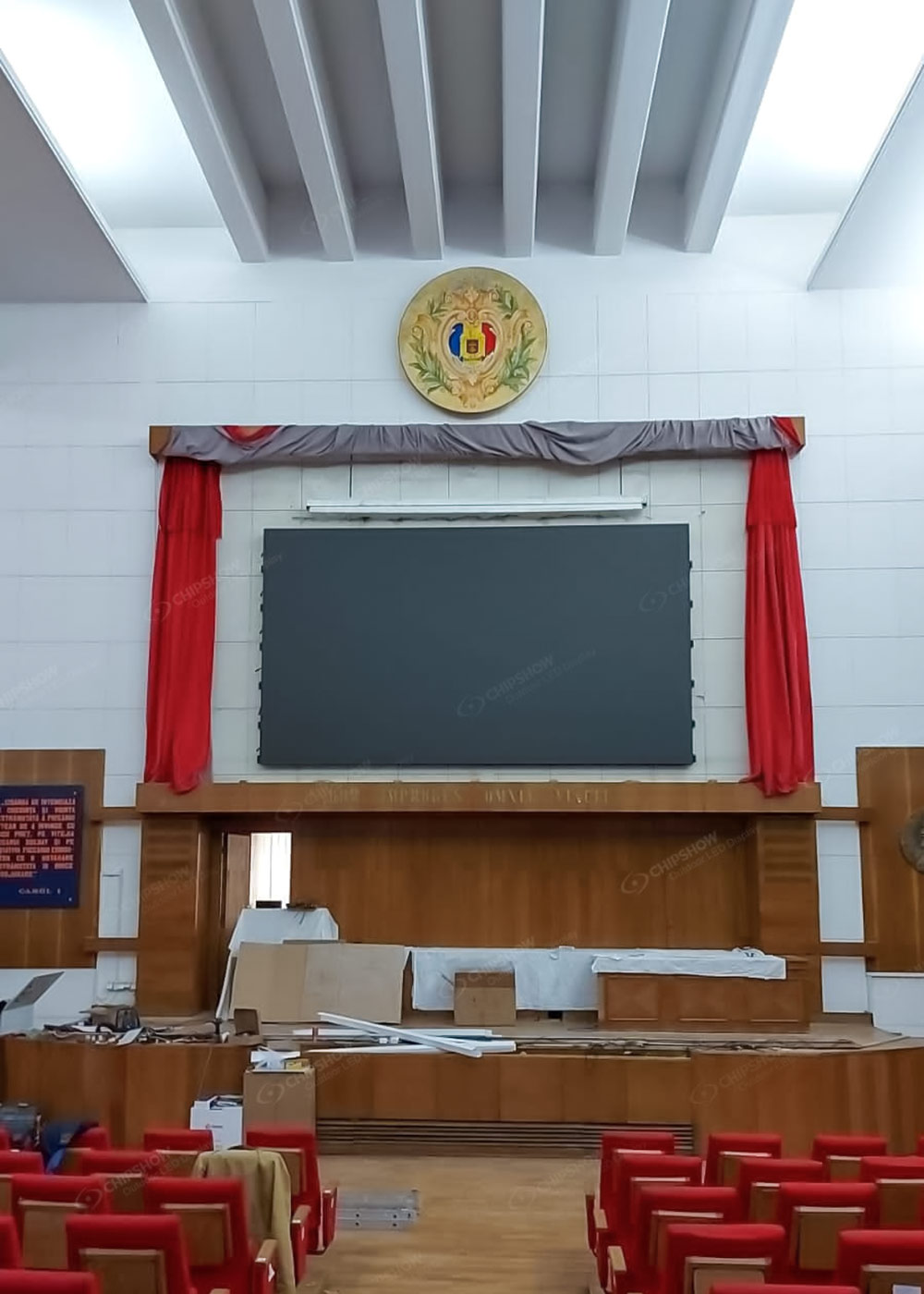 Dark screen above a wooden stage with red curtains, a crest and rows of red seats.