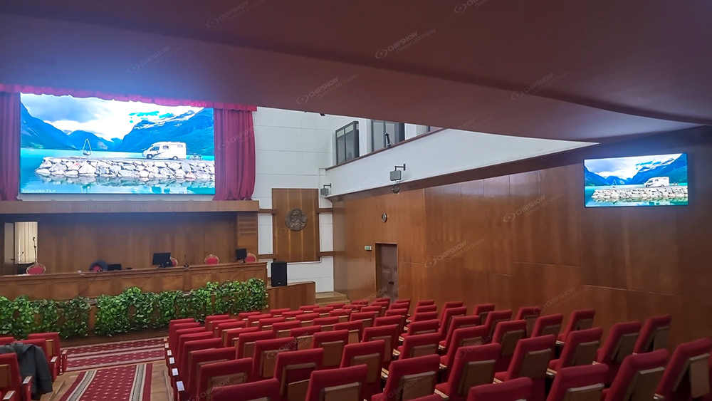 Hall with rows of red seats and two screens showing the same mountain landscape.