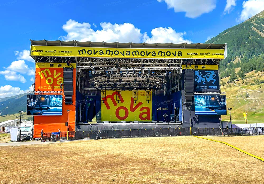 Outdoor festival stage with a yellow header, side screens and mountains behind it.