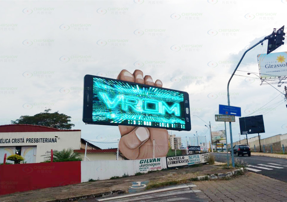 Creative outdoor LED billboard shaped like a giant hand holding a smartphone, displaying the word "VROM" in bright blue