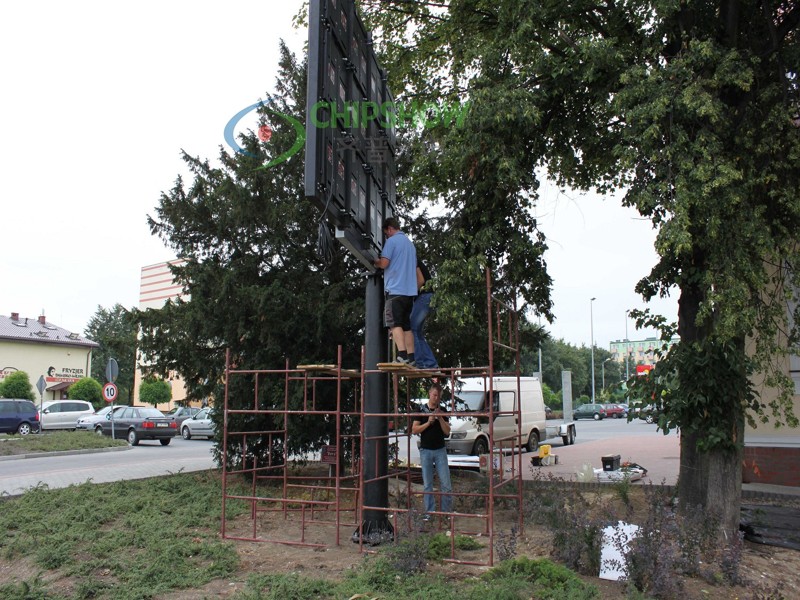 Workers on scaffolding installing or repairing a large roadside billboard beside trees and a street