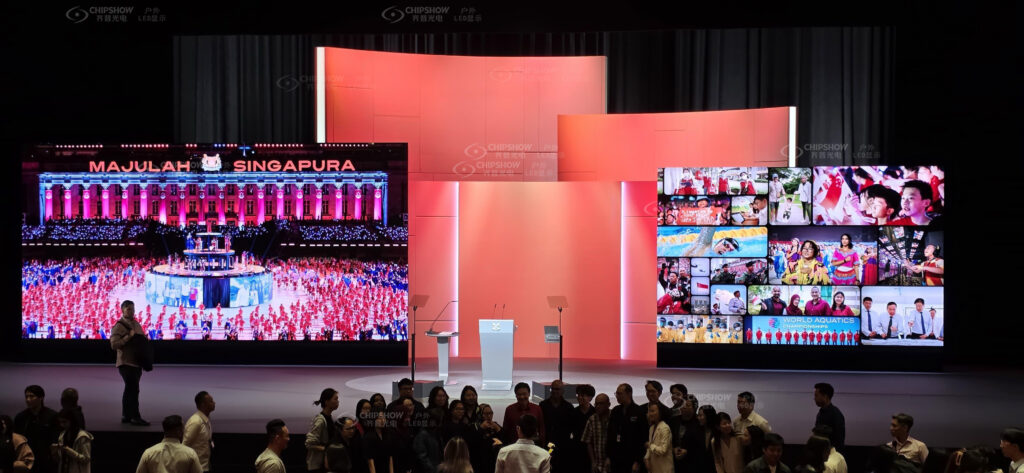 Large LED display wall with vibrant red and pink visuals on a dark conference or event stage with silhouetted audience