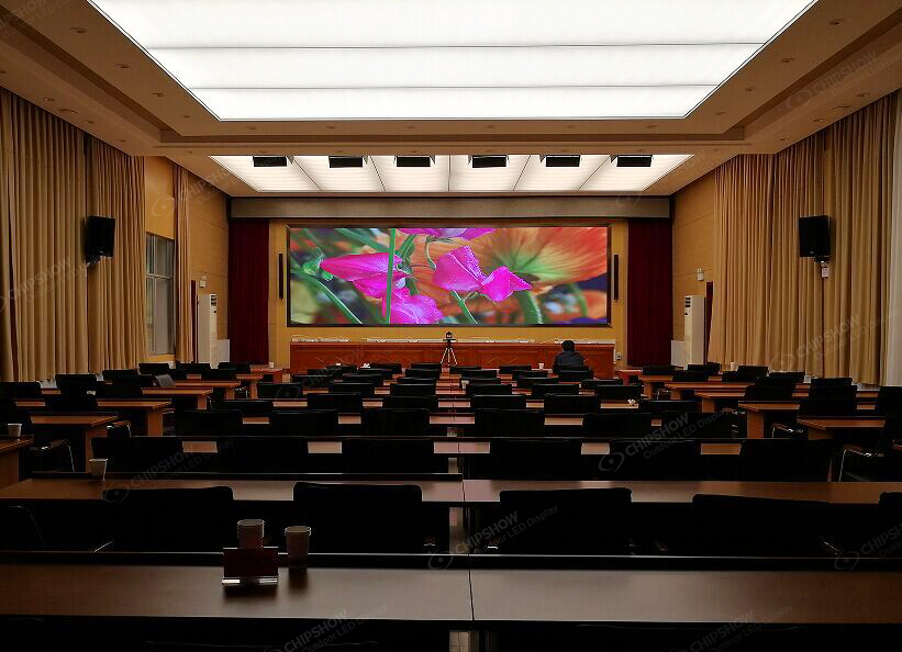 A large LED video wall displaying colorful graphics in a formal conference room with rows of empty desks.
