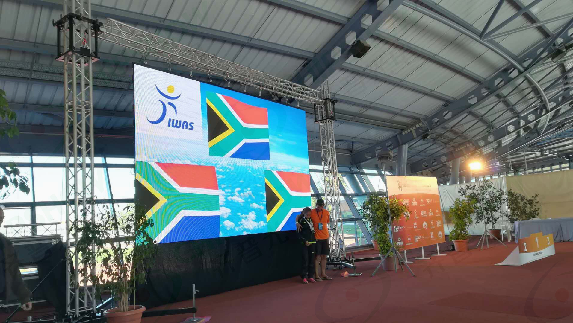 Large indoor LED display screen showing a colorful graphic at an outdoor event stage with audience and canopy structure