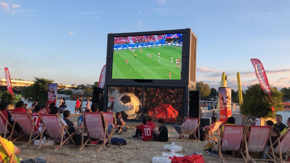 Outdoor LED sports display showing a live football match to fans seated in deck chairs on a riverside beach at sunset